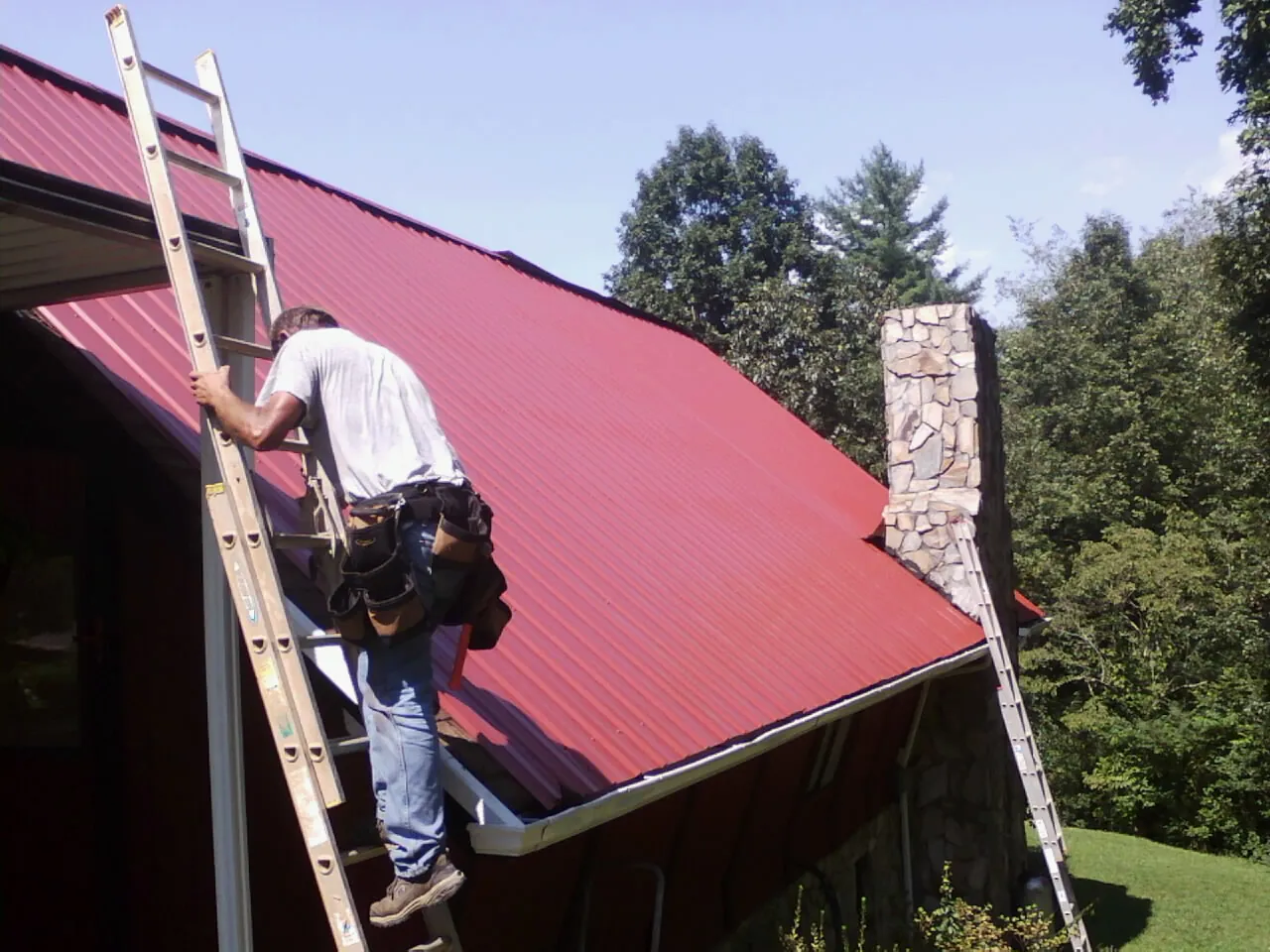 Roofer on a ladder beside a red metal roof and stone chimney — Soffit & Fascia Repair by Roofing Brothers in Westernville